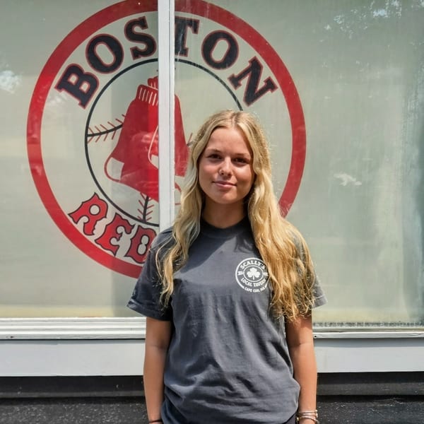Blonde woman in a gray Scally's Local Tavern t-shirt stands in front of a Boston Red Sox logo.