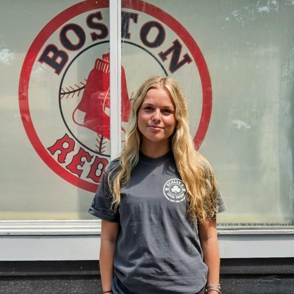 Blonde woman in a gray Scally's Local Tavern t-shirt stands in front of a Boston Red Sox logo.