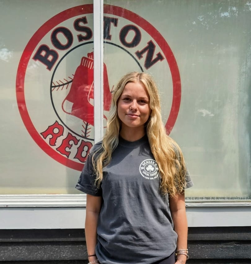 Blonde woman in a gray Scally's Local Tavern t-shirt stands in front of a Boston Red Sox logo.