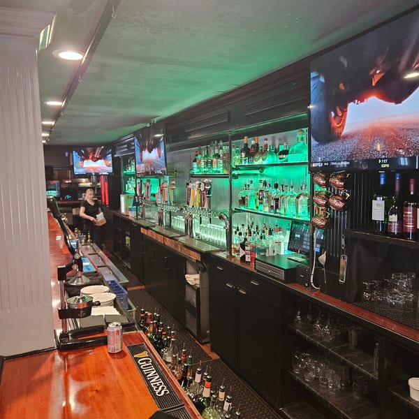 A long, well-stocked bar with green backlighting, multiple beer taps, and TVs showing sports.