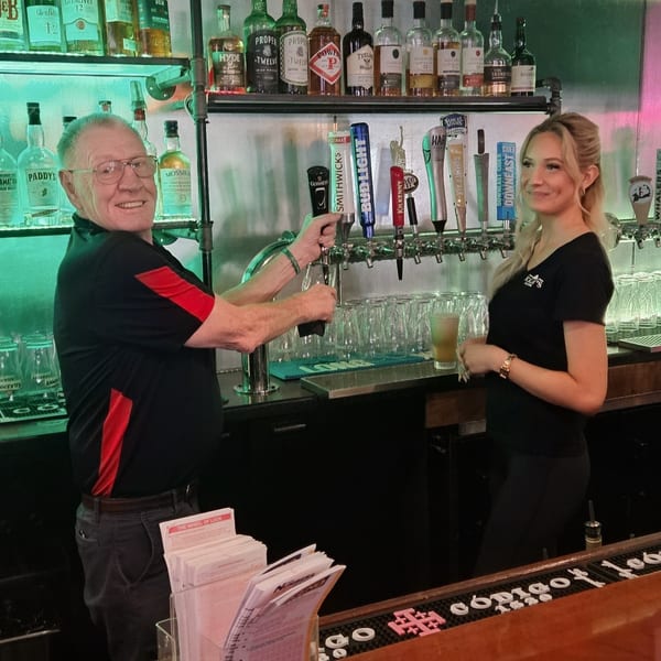Two bartenders, a man and a woman, smiling behind a bar with beer taps and liquor bottles.