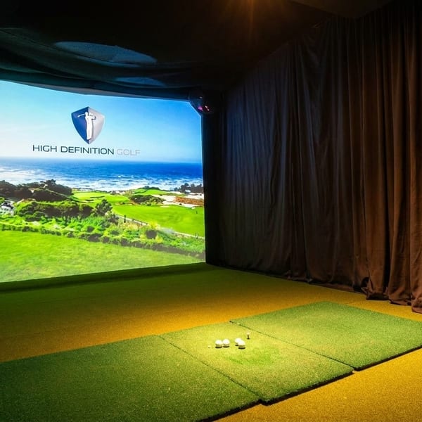 Indoor golf simulator with a projected image of a coastal golf course and golf balls on artificial turf.