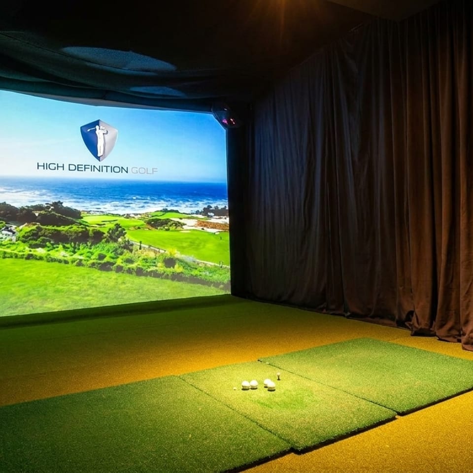 Indoor golf simulator with a projected image of a coastal golf course and golf balls on artificial turf.
