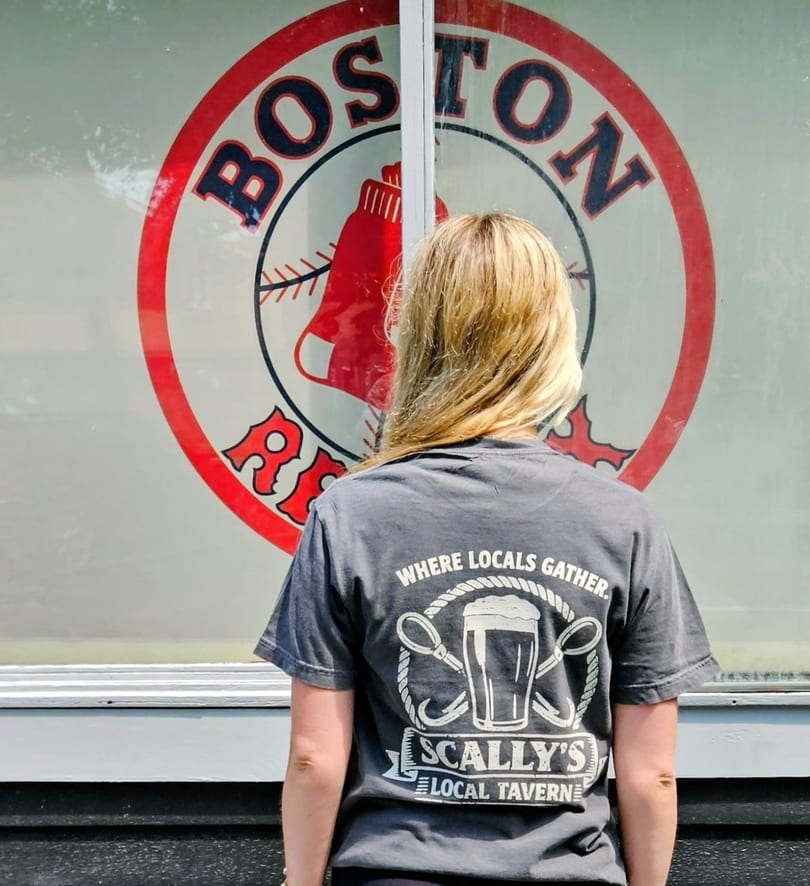 Blonde woman in a Scally's Local Tavern t-shirt looking at a Boston Red Sox logo.