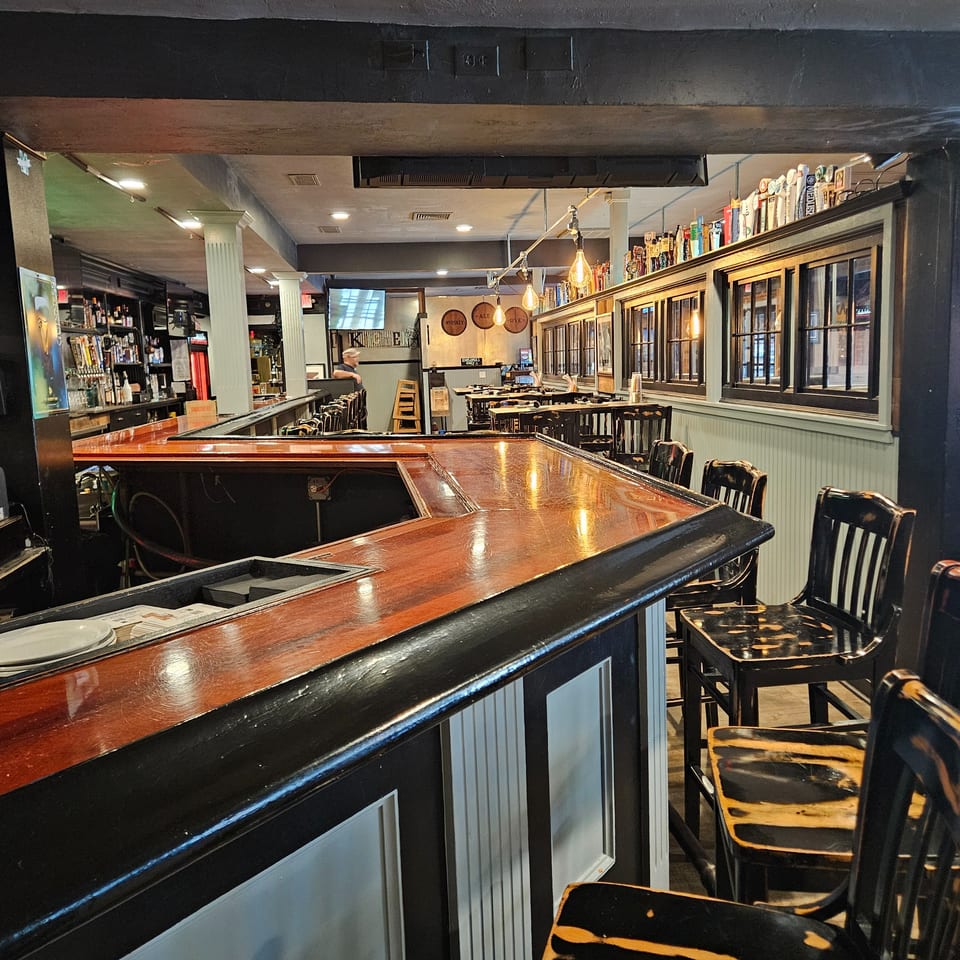 A long, polished wooden bar with black trim and bar stools in a dimly lit restaurant.