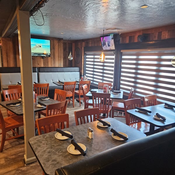 Empty restaurant dining room with wooden chairs, dark tables, booth seating, and TVs on wood-paneled walls.