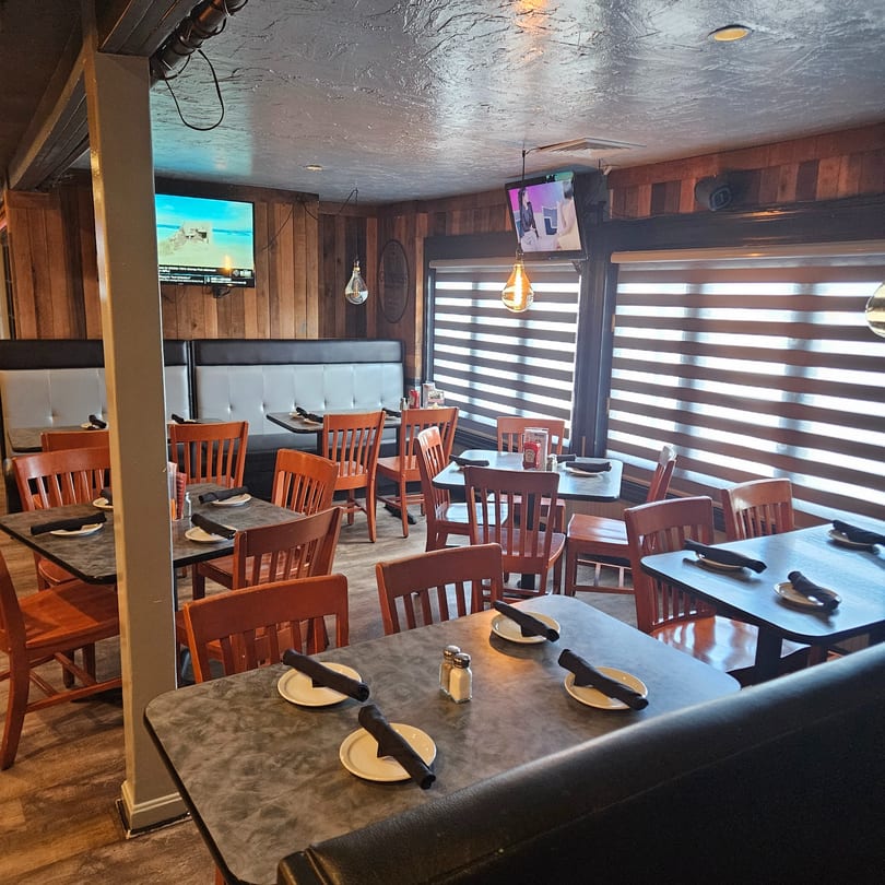 Empty restaurant dining room with wooden chairs, dark tables, booth seating, and TVs on wood-paneled walls.