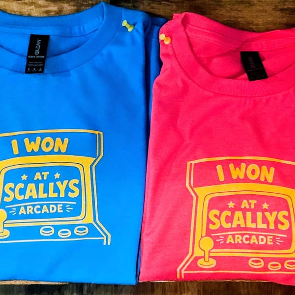 Two t-shirts, one blue and one pink, with a yellow "I WON AT SCALLY'S ARCADE" design.