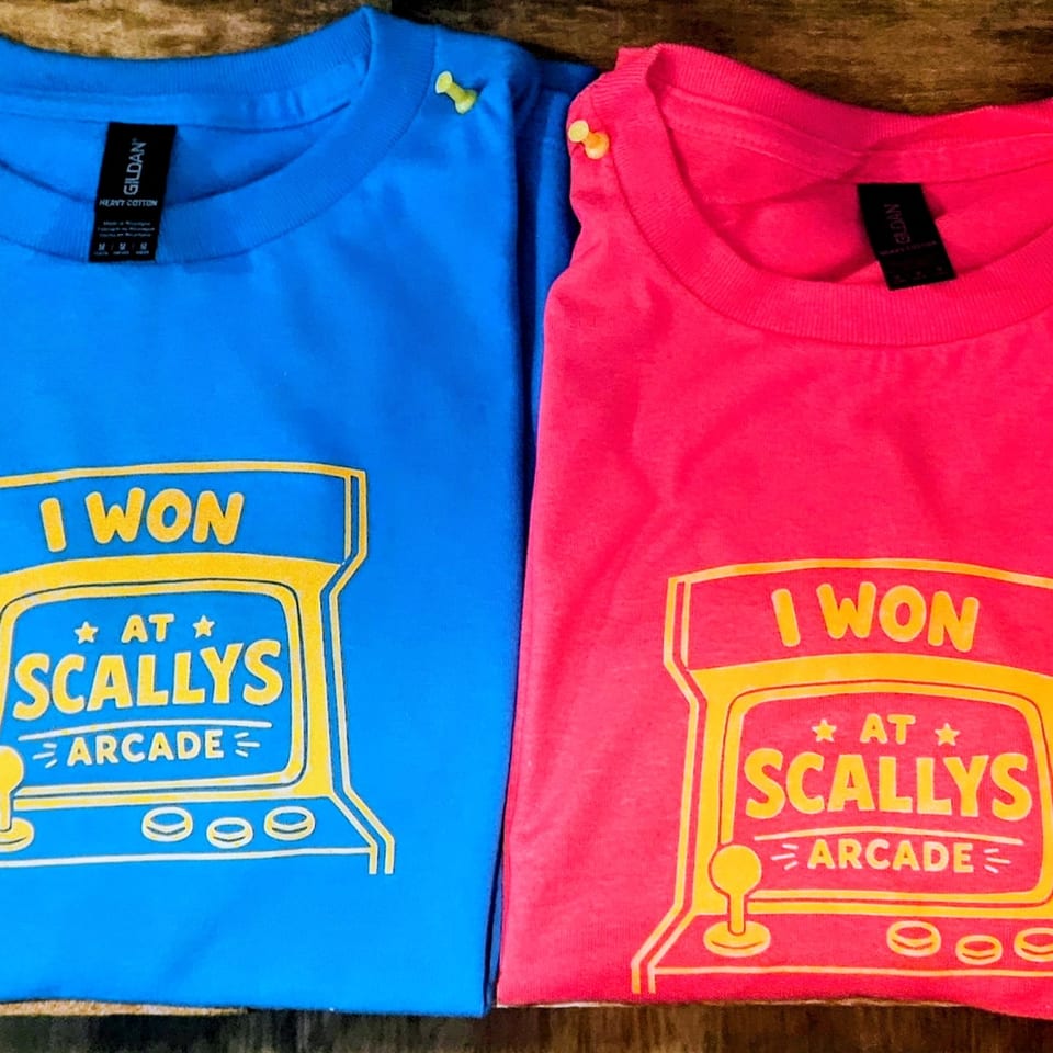 Two t-shirts, one blue and one pink, with a yellow "I WON AT SCALLY'S ARCADE" design.