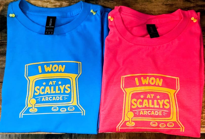 Two t-shirts, one blue and one pink, with a yellow "I WON AT SCALLY'S ARCADE" design.