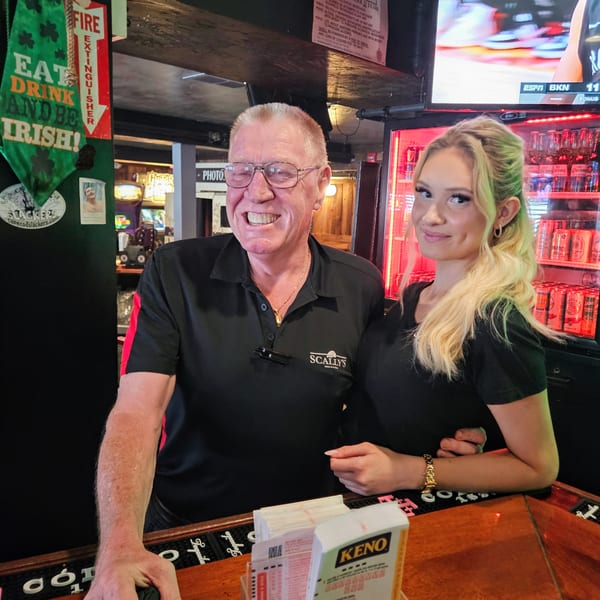 A smiling man and woman, both wearing black shirts with "SCALLY'S" logo, stand behind a bar.