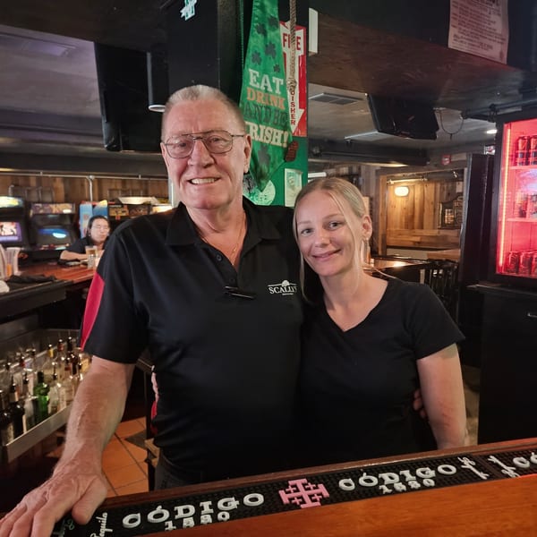 Two smiling bartenders, an older man and a younger woman, stand behind a bar with bottles and an Irish flag.