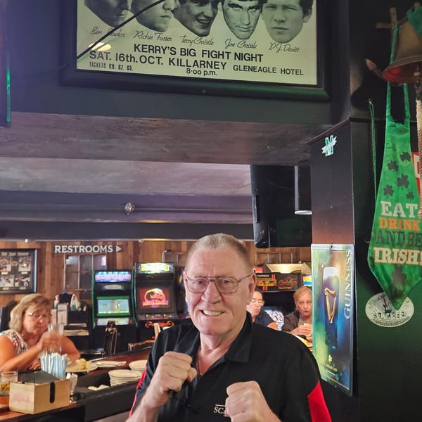 A man in a black and red shirt poses with fists up in a bar with arcade games and an Irish boxing poster.
