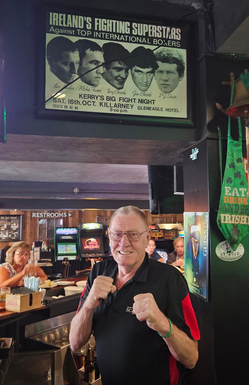 A man in a black and red shirt poses with fists up in a bar with arcade games and an Irish boxing poster.