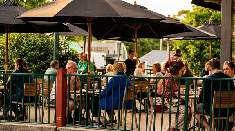 Patrons seated at outdoor tables under black umbrellas, enjoying a sunny day at a restaurant.